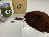 Green tumbler with an eye design next to a bag labeled 'DARK ARTS' and a pile of ground coffee on a white surface.
