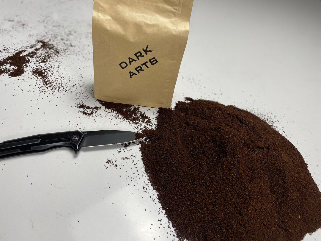 Brown paper bag labeled 'Dark Arts' standing beside a mound of ground coffee and a black knife on a white surface.